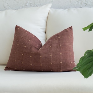 May include: A rectangular, burgundy-colored pillow with a textured woven pattern and small, square, tan accents. The pillow is placed on a white surface, with white pillows in the background. A green plant is visible on the right side.