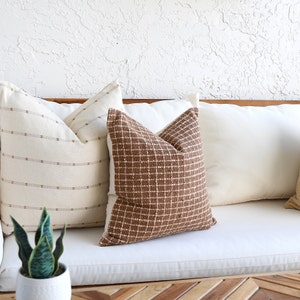 May include: Three throw pillows on a white couch. The pillows are brown, white, and beige with a geometric pattern. The couch is made of a white fabric and has a wooden frame.