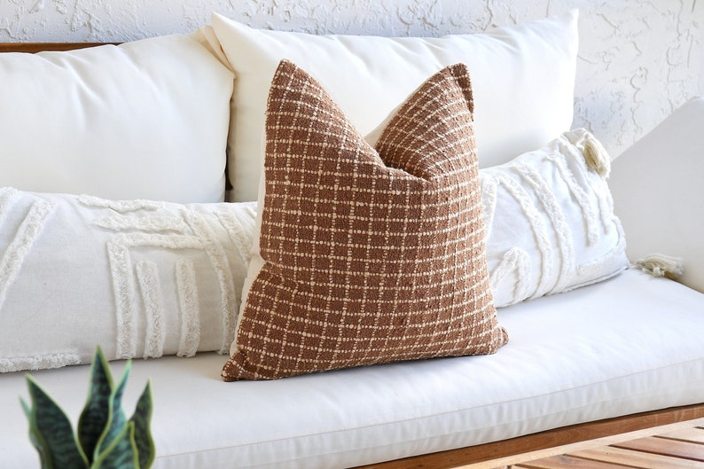 May include: A brown and white checkered throw pillow with a white fringe trim sits on a white couch with other white pillows.