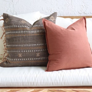 May include: Two decorative throw pillows on a white couch. One pillow is brown with a geometric pattern and fringe trim. The other pillow is solid terracotta.