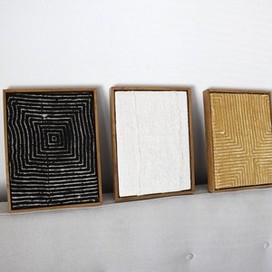 Pode incluir: Três obras de arte abstratas emolduradas. A primeira apresenta um padrão quadrado em preto e branco, a segunda uma tela branca texturizada e a terceira um design geométrico dourado. Todas estão em molduras de madeira, criando um conjunto.