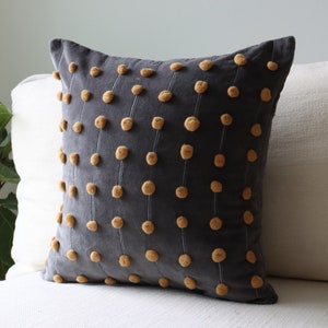 May include: A dark grey cushion featuring a pattern of vertical lines with small, round, mustard-coloured embellishments. The square cushion sits on a white sofa. A green plant is visible in the background.