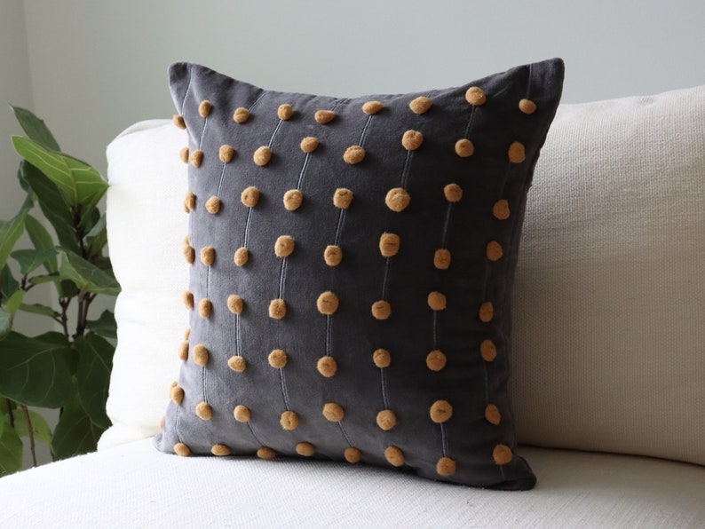 May include: A dark grey cushion featuring a pattern of vertical lines with small, round, mustard-coloured embellishments. The square cushion sits on a white sofa. A green plant is visible in the background.