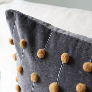 May include: A grey cushion with mustard-coloured pom-poms and white stitching. The cushion is made of a soft, velvety material. The design is modern and adds a touch of texture. It is placed against a white background.