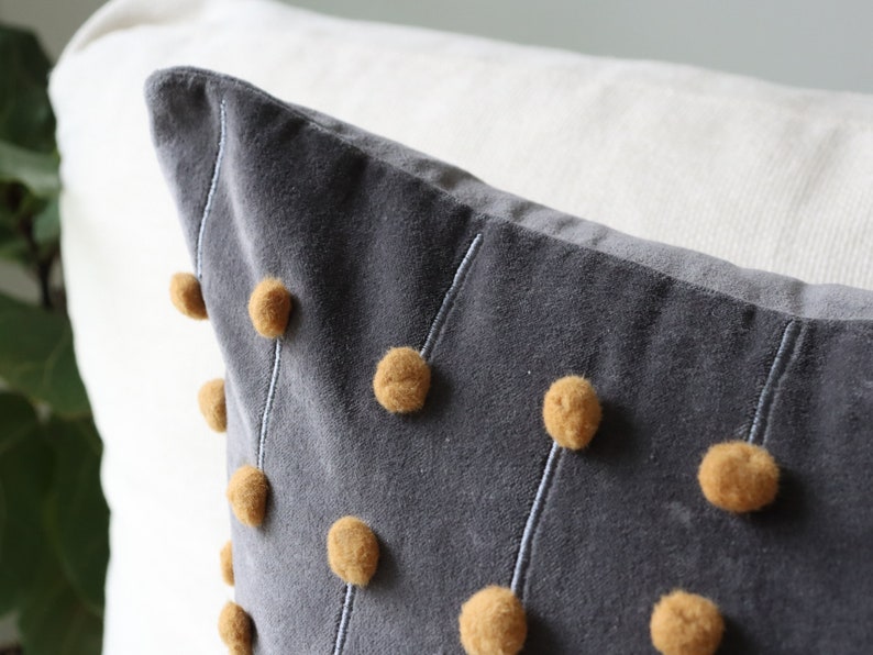 May include: A grey cushion with mustard-coloured pom-poms and white stitching. The cushion is made of a soft, velvety material. The design is modern and adds a touch of texture. It is placed against a white background.