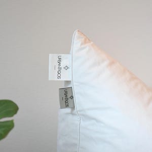 May include: Close-up of a white pillow with two tags attached. The tags are rectangular with the text "URBANITROCS PARIS" and a small geometric design. The pillow is against a white background.
