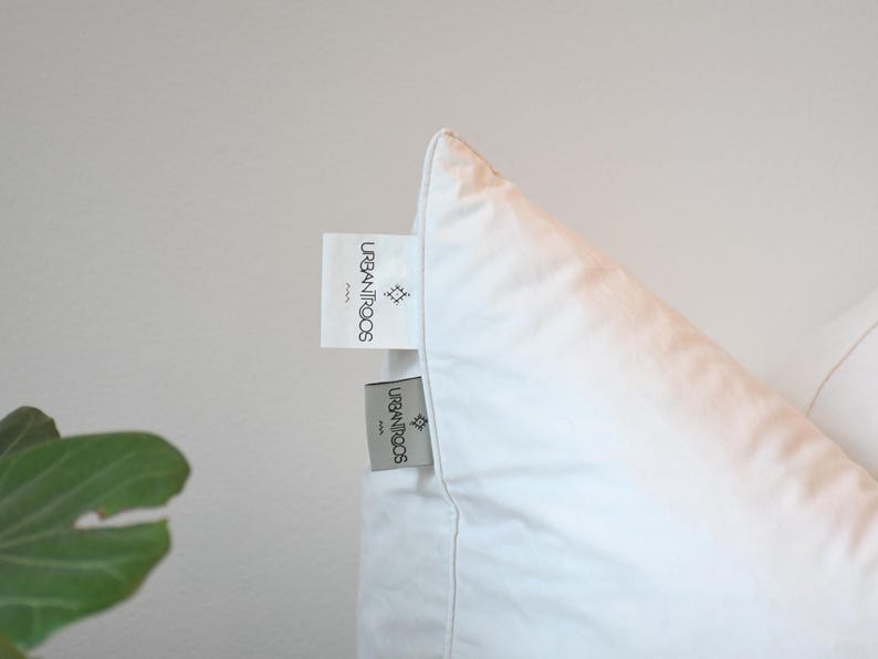 May include: Close-up of a white pillow with two tags attached. The tags are rectangular with the text "URBANITROCS PARIS" and a small geometric design. The pillow is against a white background.