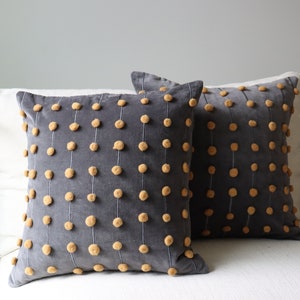 May include: Two square grey decorative cushions with vertical rows of small, tan pom-poms. The cushions are on a white surface, and the background is a light grey wall. The cushions have a soft, textured appearance.