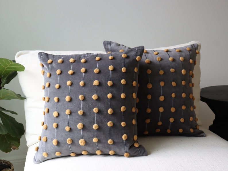 May include: Two square grey decorative cushions with vertical rows of small, tan pom-poms. The cushions are on a white surface, and the background is a light grey wall. The cushions have a soft, textured appearance.