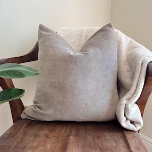May include: A large, taupe-colored pillow with a soft, textured appearance rests on a wooden chair. A cream-colored blanket is draped over the chair's back. The pillow is the focal point, suggesting comfort and home decor.