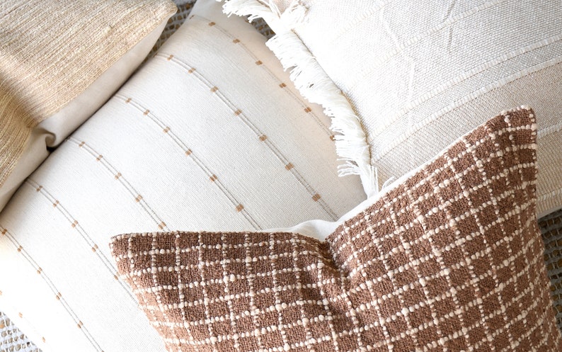 May include: Three decorative throw pillows with different textures and patterns. The pillows are made of a soft, woven fabric. The first pillow is a light brown with a woven texture. The second pillow is white with thin brown stripes. The third pillow is brown with a woven grid pattern.