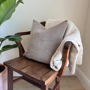 May include: A taupe-colored pillow rests on a vintage wooden chair, with a cream-colored blanket draped over the back. The chair has a dark wood finish. A potted plant with large green leaves is in the background.