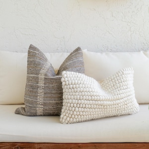 May include: A sofa with three decorative pillows. One pillow is gray with a white stripe design, another is white with a textured, bobble design, and the third is a plain white pillow. The sofa has a wooden frame.