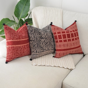 May include: Three square throw pillows featuring intricate patterns in red, black, and cream. Each pillow is finished with small black pom-poms at the four corners. The designs include geometric shapes, lines, and swirls printed on the fabric.