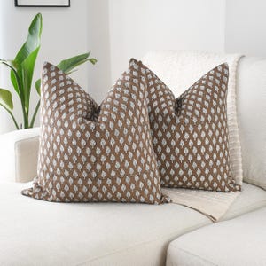 May include: Two brown throw cushions with a white leaf pattern on a white sofa. The cushions are square and have a textured appearance. A white blanket is behind the cushions, and a green plant is in the background.