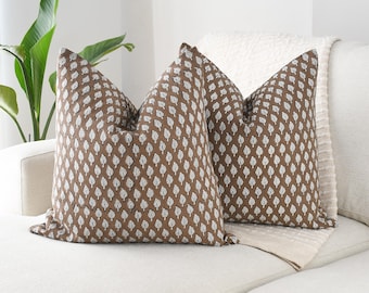 Può includere: Due cuscini decorativi marroni con un motivo a foglie bianche su un divano bianco. I cuscini quadrati hanno un aspetto strutturato. Una coperta bianca è dietro i cuscini e una pianta verde è visibile sullo sfondo.