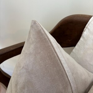 May include: Close-up of a light taupe-colored pillow with a pointed top. The pillow is made of a soft, velvety material. The pillow is resting on a dark brown wooden chair.