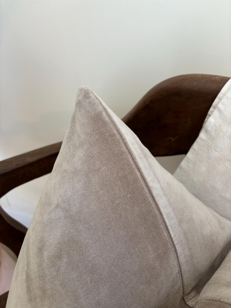 May include: Close-up of a light taupe-colored pillow with a pointed top. The pillow is made of a soft, velvety material. The pillow is resting on a dark brown wooden chair.