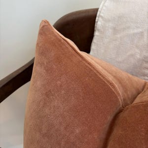May include: Close-up of a terracotta-colored velvet pillow. The pillow is on a dark wooden chair. The pillow has a soft, plush texture and a stitched seam along the edge. A light-colored pillow is partially visible in the background.