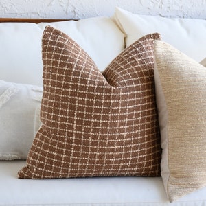 May include: A brown and white patterned throw pillow with a grid design. The pillow is on a white couch.
