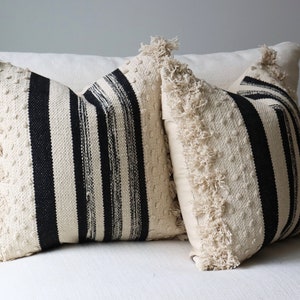 May include: Two decorative throw pillows with a cream-colored background and black stripes. The pillows have a textured design and fringe trim along the edges. They are placed on a white sofa.