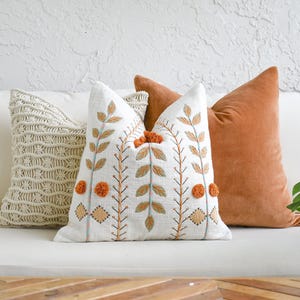 Pode incluir: Uma coleção de almofadas decorativas em várias texturas e cores. As almofadas incluem uma almofada branca, uma almofada de macramé creme, uma almofada branca com folhas bordadas e pompons laranja e uma almofada de veludo enferrujado.