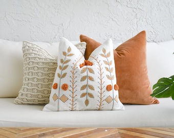 Puede incluir: Una colección de cojines decorativos en varias texturas y colores. Los cojines incluyen un cojín blanco, un cojín de macramé color crema, un cojín blanco con hojas bordadas y pompones naranjas, y un cojín de terciopelo color óxido.