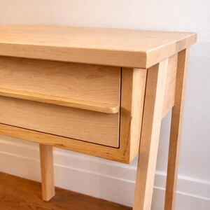 Pu&ograve; includere: Un tavolino in legno chiaro con un singolo cassetto. Il tavolo ha un piano rettangolare e quattro gambe angolate. Il cassetto ha un design semplice e minimalista con una piccola maniglia. La venatura del legno &egrave; visibile.