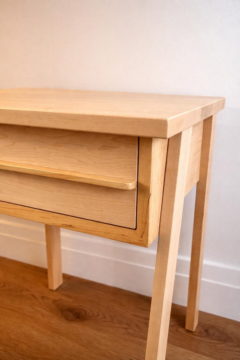 Pu&ograve; includere: Un tavolino in legno chiaro con un singolo cassetto. Il tavolo ha un piano rettangolare e quattro gambe angolate. Il cassetto ha un design semplice e minimalista con una piccola maniglia. La venatura del legno &egrave; visibile.