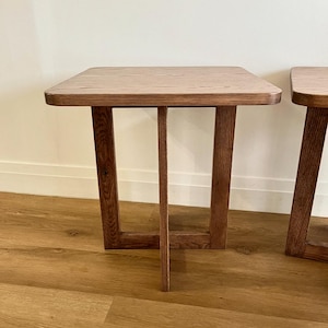 May include: Two wooden side tables with square tops and angled legs. The tables are made of light brown wood and have a simple, modern design. The tables are placed on a light brown wooden floor.