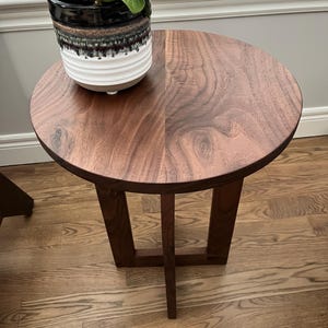 Op de afbeelding: Een ronde bijzettafel van walnoot hout met een eenvoudig, modern ontwerp. De tafel heeft een donkerbruine afwerking en een enkel, open rek. Een plant in een pot staat op het tafelblad.