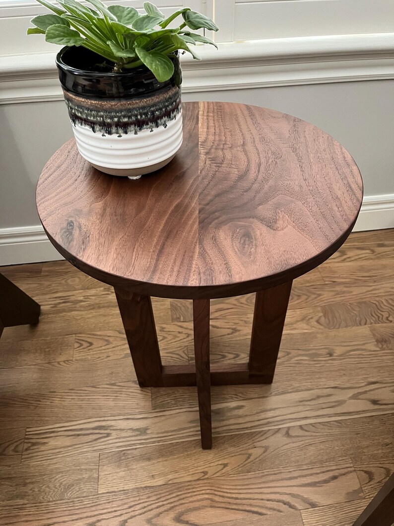 Op de afbeelding: Een ronde bijzettafel van walnoot hout met een eenvoudig, modern ontwerp. De tafel heeft een donkerbruine afwerking en een enkel, open rek. Een plant in een pot staat op het tafelblad.