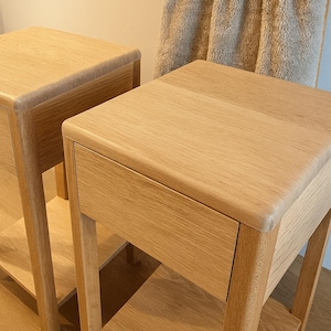 Puede incluir: Dos mesitas de noche de roble claro con un caj&oacute;n y un estante inferior. Las mesas tienen un dise&ntilde;o sencillo y moderno con bordes redondeados. Una manta beige cuelga en el fondo.