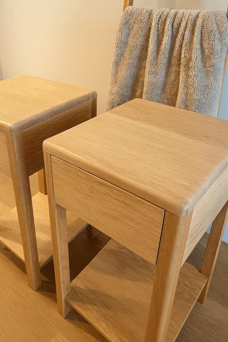 Puede incluir: Dos mesitas de noche de roble claro con un caj&oacute;n y un estante inferior. Las mesas tienen un dise&ntilde;o sencillo y moderno con bordes redondeados. Una manta beige cuelga en el fondo.