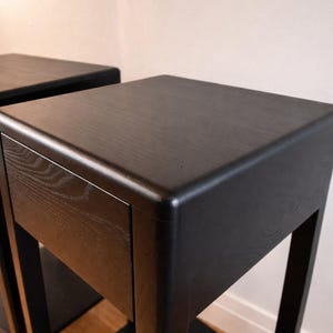 Puede incluir: Dos mesas auxiliares de madera negra con un dise&ntilde;o minimalista. Cada mesa tiene una parte superior cuadrada, un caj&oacute;n y un estante inferior. Las mesas est&aacute;n hechas de madera oscura y tienen un acabado liso y mate. Las patas son cuadradas y robustas.