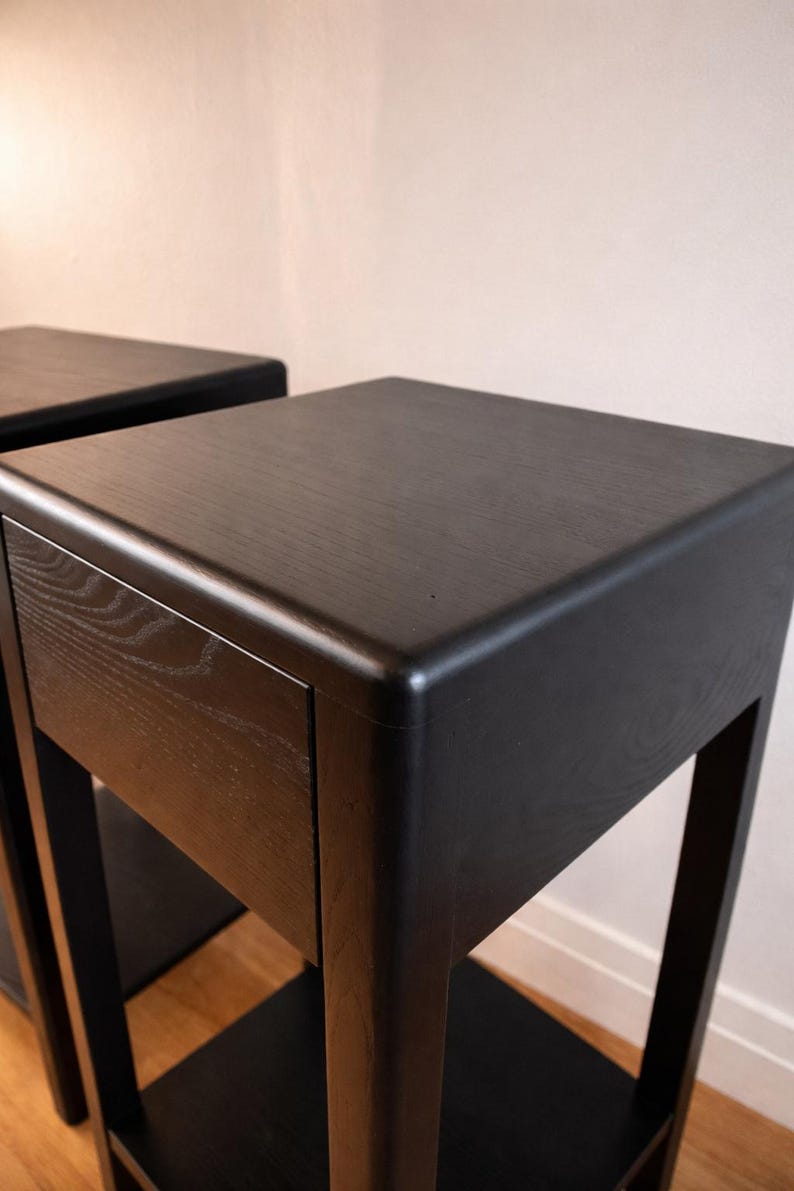 Puede incluir: Dos mesas auxiliares de madera negra con un dise&ntilde;o minimalista. Cada mesa tiene una parte superior cuadrada, un caj&oacute;n y un estante inferior. Las mesas est&aacute;n hechas de madera oscura y tienen un acabado liso y mate. Las patas son cuadradas y robustas.