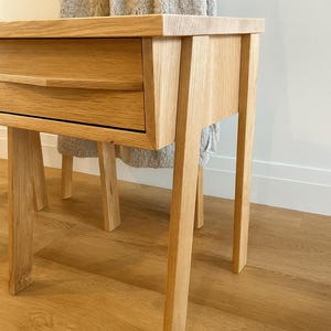 Pu&ograve; includere: Un tavolino in legno di rovere chiaro con un cassetto. Il tavolo ha un piano rettangolare e quattro gambe dritte. Il cassetto ha una maniglia curva. Il tavolo &egrave; appoggiato su un pavimento in legno marrone chiaro.