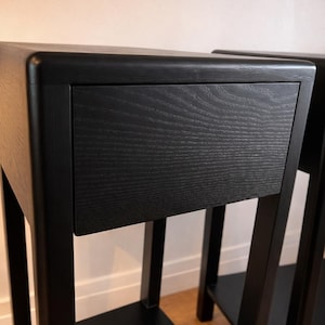 Puede incluir: Dos mesitas de noche de madera negra con un caj&oacute;n. Las mesitas tienen un dise&ntilde;o minimalista con una parte superior rectangular y un estante inferior. La veta de la madera es visible en los frentes de los cajones. Las patas son cuadradas y robustas.