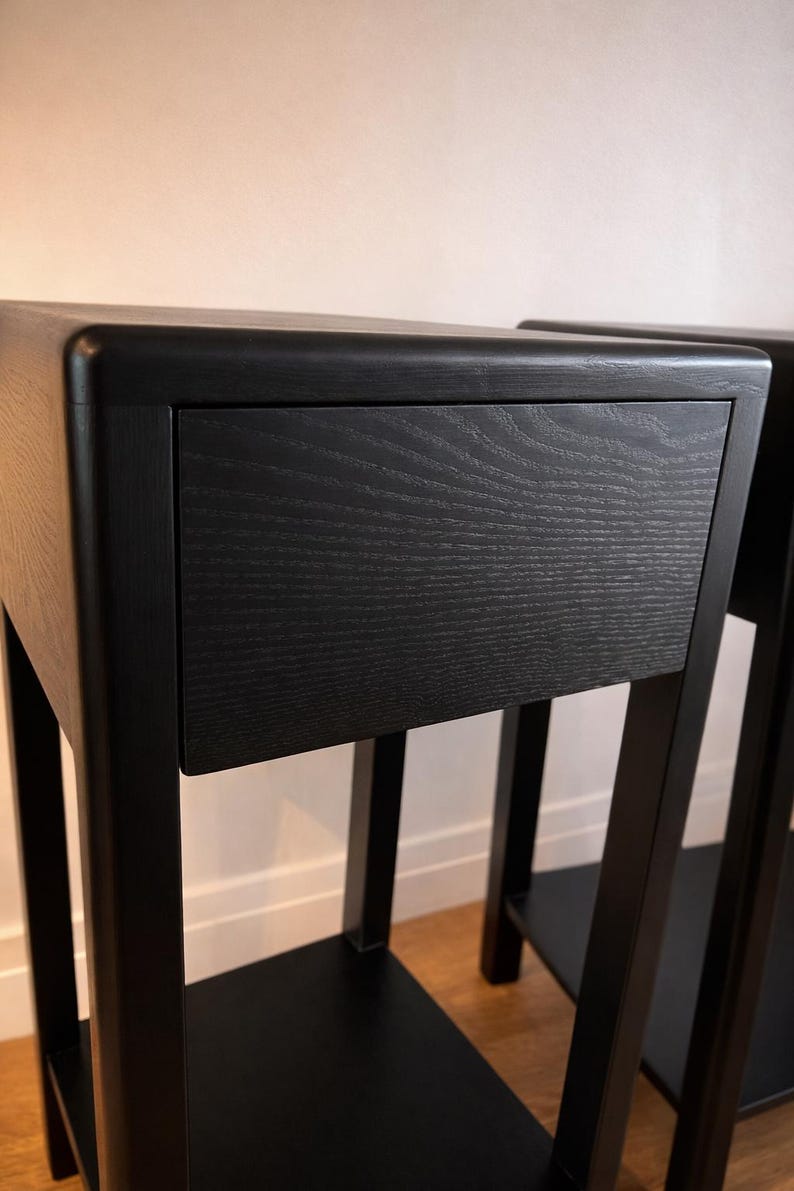 Puede incluir: Dos mesitas de noche de madera negra con un caj&oacute;n. Las mesitas tienen un dise&ntilde;o minimalista con una parte superior rectangular y un estante inferior. La veta de la madera es visible en los frentes de los cajones. Las patas son cuadradas y robustas.
