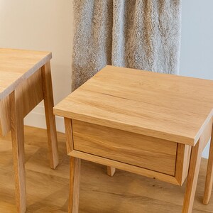 Pu&ograve; includere: Due comodini in legno di rovere chiaro con un cassetto, con finitura a venatura naturale. I tavoli hanno piani quadrati e quattro gambe dritte. Una soffice coperta colorata &egrave; appesa sullo sfondo.