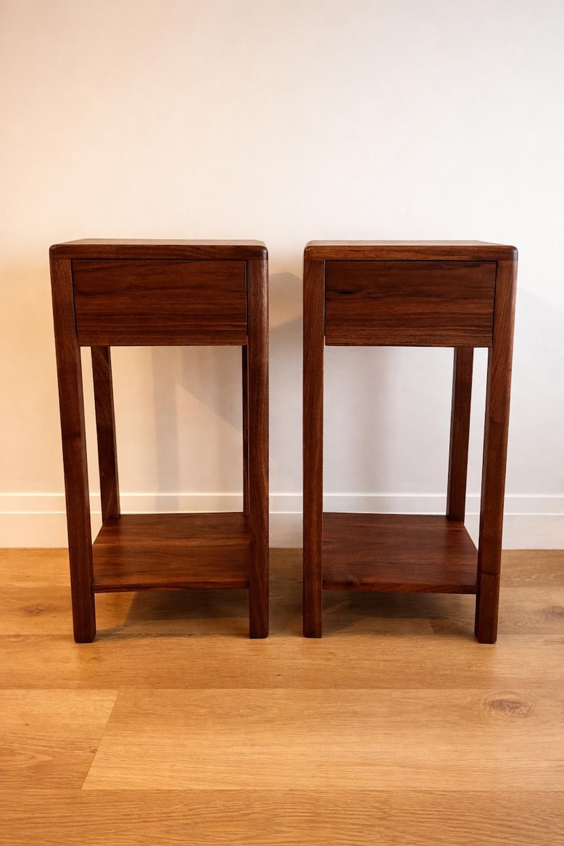 Puede incluir: Dos mesas auxiliares de madera a juego. Cada mesa tiene un caj&oacute;n, un estante inferior y cuatro patas. Las mesas son de madera marr&oacute;n oscuro y est&aacute;n colocadas contra una pared blanca y un suelo de madera marr&oacute;n claro.