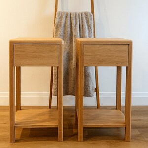 Puede incluir: Dos mesitas de noche de madera a juego con cajones y estantes inferiores. Las mesas son de madera de color claro y tienen un dise&ntilde;o sencillo y moderno. Una escalera de madera con una manta gris est&aacute; en el fondo.