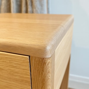 Puede incluir: Primer plano de una mesita de noche de madera de roble claro. La mesita de noche presenta un borde redondeado en la parte superior y un solo caj&oacute;n. La veta de la madera es visible y la pieza tiene un tono natural y c&aacute;lido. El dise&ntilde;o es minimalista y moderno.
