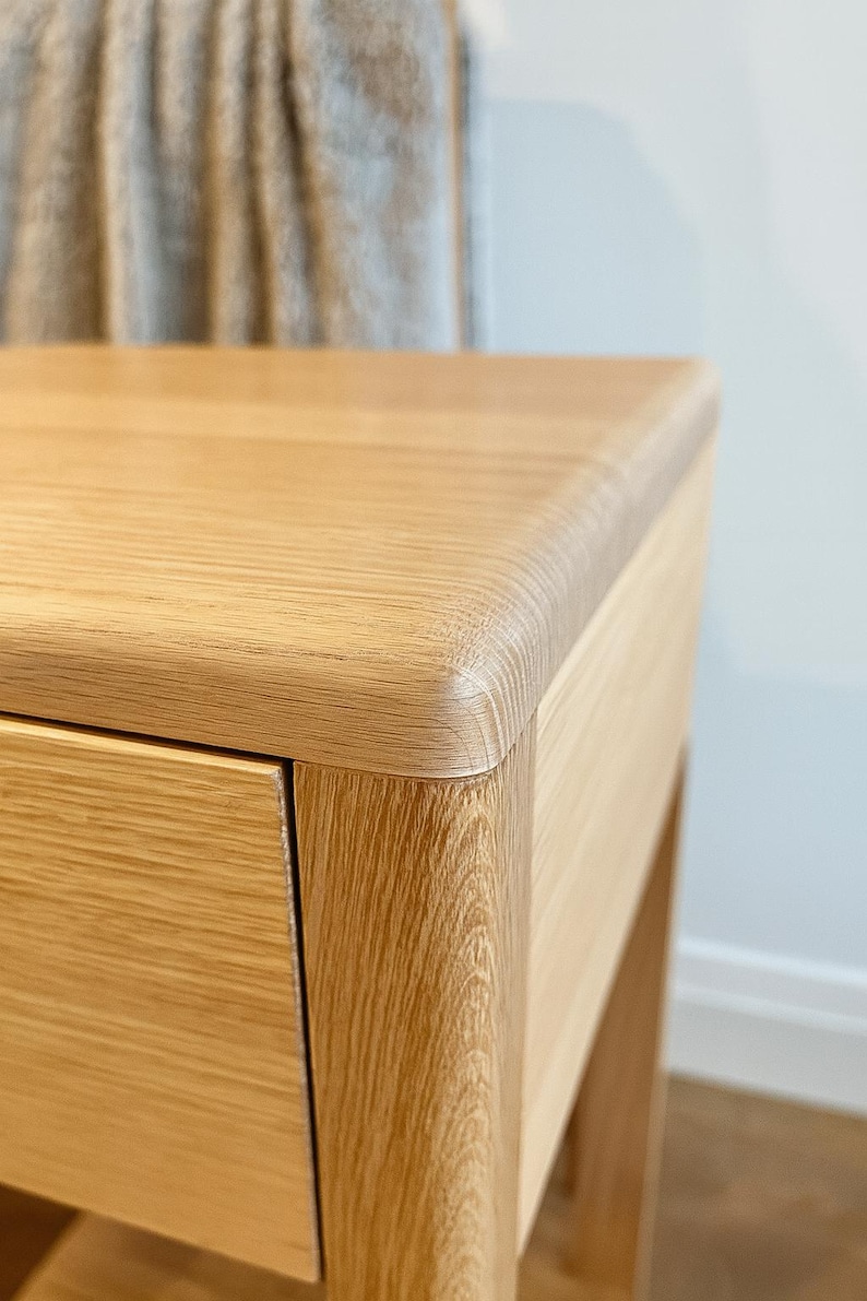 Puede incluir: Primer plano de una mesita de noche de madera de roble claro. La mesita de noche presenta un borde redondeado en la parte superior y un solo caj&oacute;n. La veta de la madera es visible y la pieza tiene un tono natural y c&aacute;lido. El dise&ntilde;o es minimalista y moderno.