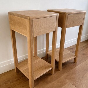 Puede incluir: Dos mesitas de noche de madera a juego. Cada una tiene una parte superior cuadrada con un caj&oacute;n, sostenida por cuatro patas. Una balda inferior est&aacute; situada entre las patas. Las mesitas est&aacute;n hechas de madera clara y se apoyan en un suelo de madera.
