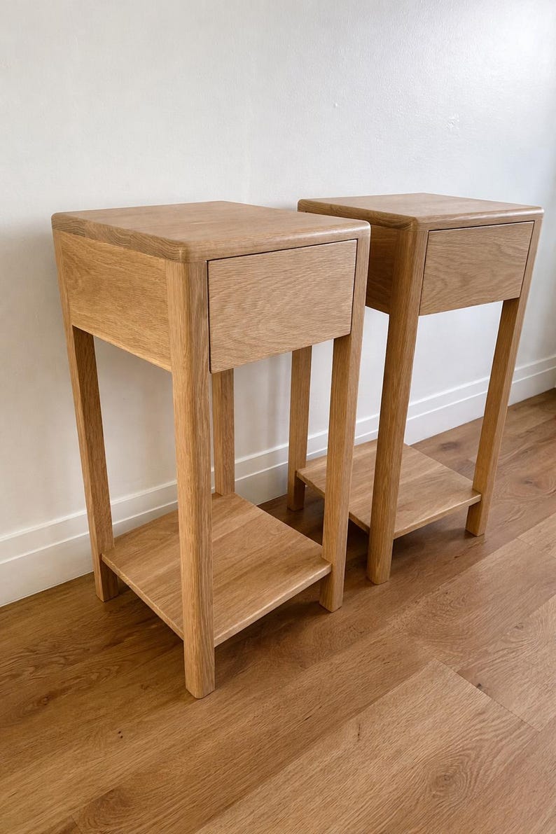 Puede incluir: Dos mesitas de noche de madera a juego. Cada una tiene una parte superior cuadrada con un caj&oacute;n, sostenida por cuatro patas. Una balda inferior est&aacute; situada entre las patas. Las mesitas est&aacute;n hechas de madera clara y se apoyan en un suelo de madera.