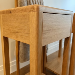 Puede incluir: Mesita de noche de roble claro con un solo caj&oacute;n. La mesa tiene esquinas redondeadas y un estante inferior. Un toallero de madera con una toalla beige est&aacute; en el fondo. La mesa est&aacute; hecha de madera maciza.