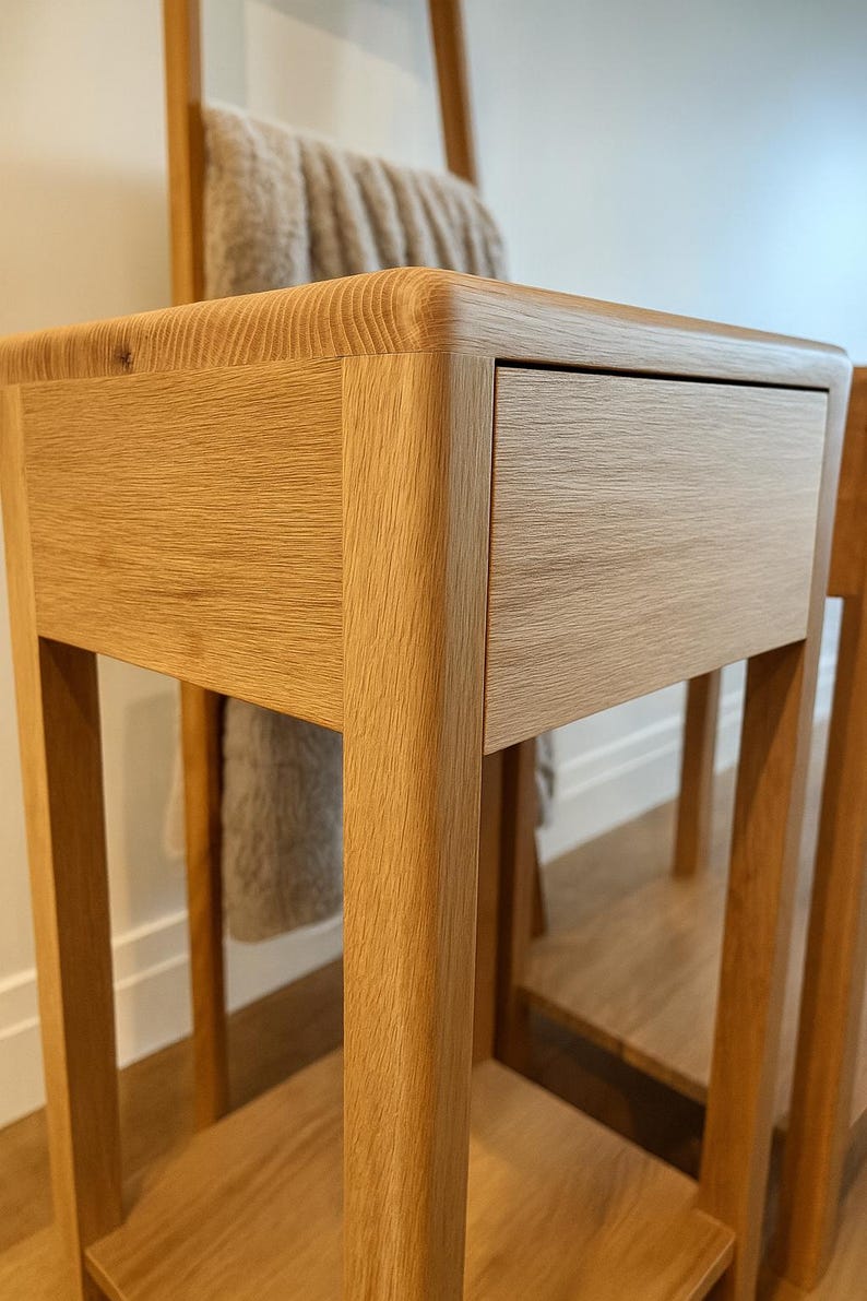Puede incluir: Mesita de noche de roble claro con un solo caj&oacute;n. La mesa tiene esquinas redondeadas y un estante inferior. Un toallero de madera con una toalla beige est&aacute; en el fondo. La mesa est&aacute; hecha de madera maciza.