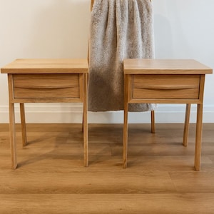 Puede incluir: Dos mesitas de noche de madera a juego, cada una con un solo cajón. Las mesitas están hechas de madera de color claro y tienen un diseño sencillo y moderno. Una escalera de madera con una manta en la parte trasera.