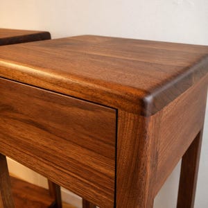 Puede incluir: Dos mesitas de noche de madera con un acabado marr&oacute;n c&aacute;lido. Cada mesa tiene un caj&oacute;n y un estante inferior. La veta de la madera es visible, dando un aspecto natural. El dise&ntilde;o es minimalista y moderno, adecuado para un dormitorio.